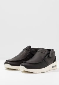 Mustat nahkaiset liukusneakersit, joissa on valkoiset kumipohjat ja tikatut yksityiskohdat, esiteltyinä vierekkäin valkoisella pinnalla.