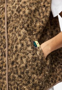 Brūns un melns teksturēts flīša vestes ar roku sānu kabatā, bēša apmale un neliela zilā un dzeltenā auduma birka uz kabatas malas.