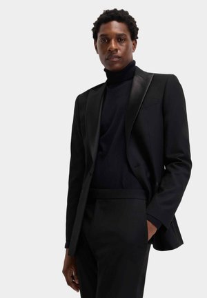 Schwarzer, maßgeschneiderter Anzug mit Satinrevers, kombiniert mit einem dunklen Rollkragenpullover. Glatte Stoffstruktur; figurbetonte Form mit klaren Linien.