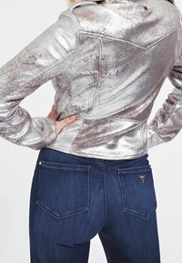 Metaliczna srebrna kurtka o fakturowanej powierzchni, z ostrym kołnierzem i zakrzywionymi szwami, połączona z ciemnoniebieskimi dżinsami z detalami kieszeni.