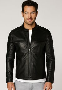 Uomo con giacca di pelle nera con zip e patch cuciti sulle spalle, camicia bianca sotto e pantaloni grigio chiaro, in piedi contro uno sfondo neutro.