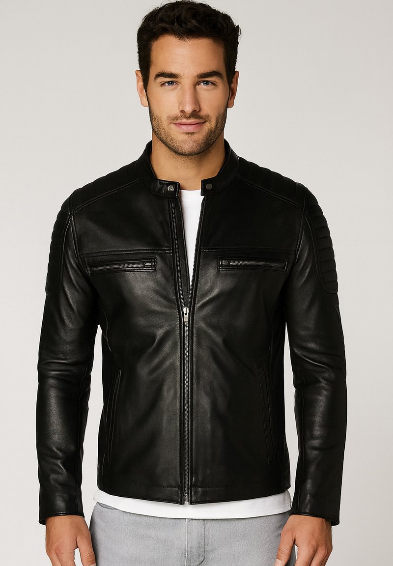Uomo con giacca di pelle nera con zip e patch cuciti sulle spalle, camicia bianca sotto e pantaloni grigio chiaro, in piedi contro uno sfondo neutro.