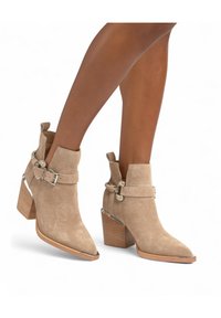 Gli stivali alla caviglia in suede beige presentano una punta affusolata, abbellimenti argentati, una fibbia decorativa e un tacco impilato con un accento argento.
