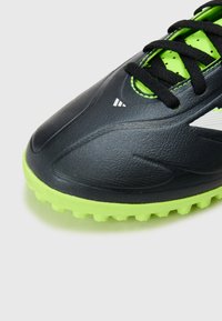 Zwarte voetbalschoen met een glanzend synthetisch bovenwerk, groene accenten, een textuur op het oppervlak en een rubberen buitenzool met korte noppen voor grip.