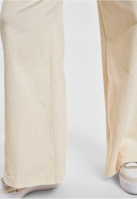 Cremfarbene Cordhosen mit weitem Beinschnitt, gerippter Textur und sichtbaren Nähdetails. Schuhe mit weißem Grund und beigen Akzenten.