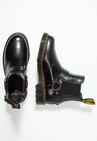 Une paire de bottes chelsea en cuir noir avec des coutures jaunes, des sangles à boucle, des panneaux latéraux élastiques et des languettes de talon avec la marque sur fond blanc.