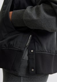 Zwarte bomberjack met geribbelde zoom, voorzien van een grijze wollen mouw. Heeft een label met "Marc O'Polo" en metalen drukknopen.