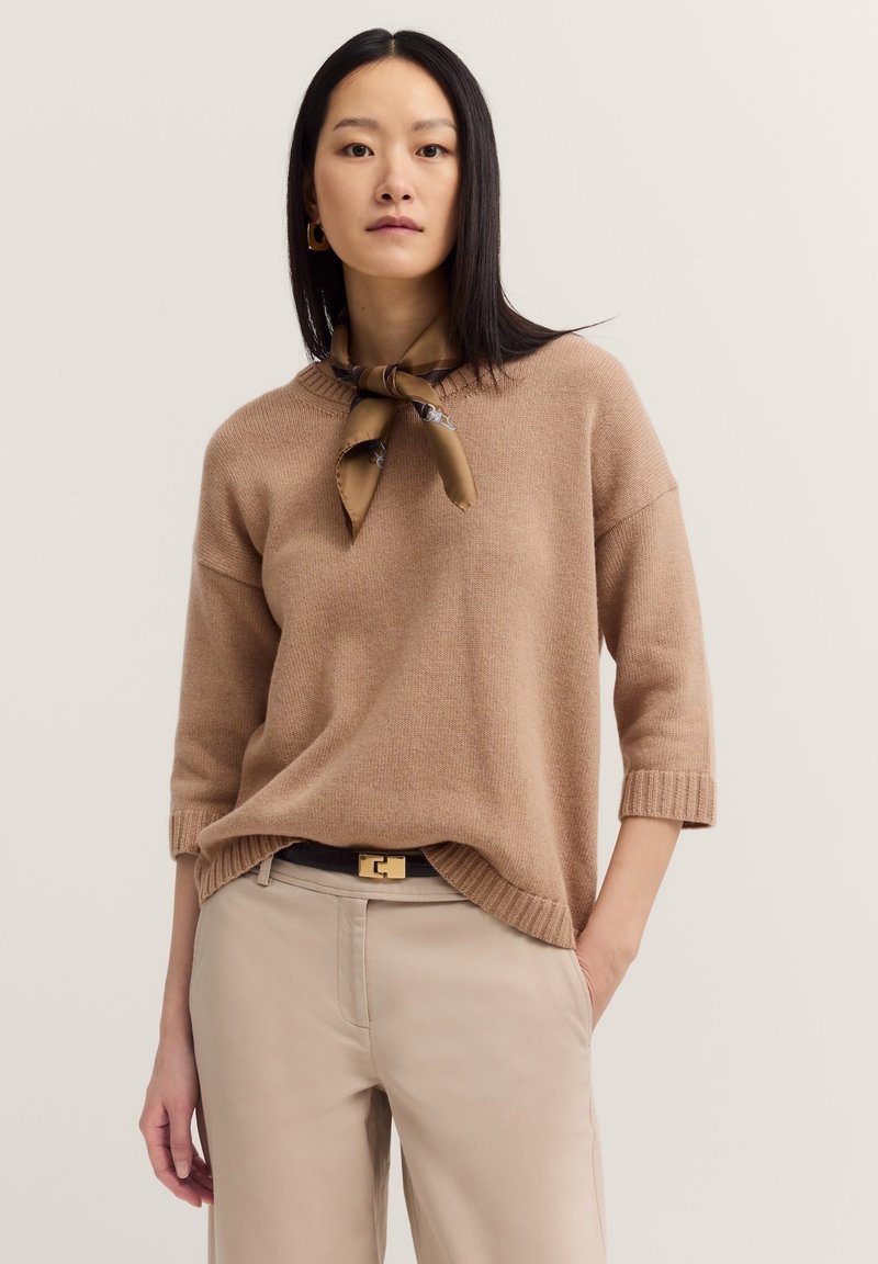 Beige Strickpullover in entspannter Passform mit ärmellangen Ärmeln. Kombiniert mit cremefarbenen Hosen und einem braunen Seidenschal, der um den Hals gebunden ist.