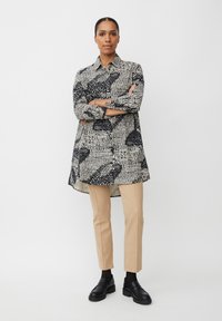 Schwarz-weiß gemustertes Hemdkleid mit langen Ärmeln, einem Kragen und einem asymmetrischen Saum, kombiniert mit beigen Slim-Hosen und schwarzen Schuhen.