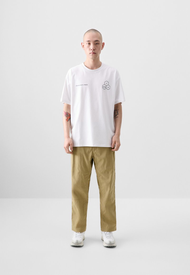 White cotton t-shirt with graphic print on the chest, paired with khaki trousers and white sneakers. Simple, relaxed fit.