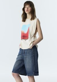 Mujer con cabello castaño hasta los hombros, ropa sin mangas color crema con gráfico rojo y azul y pantalones cortos de mezclilla sueltos hasta la rodilla, manos en los bolsillos.