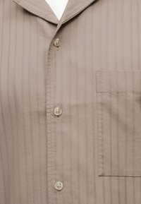 Μπεζ πουκάμισο με κουμπιά, λεπτές κάθετες ρίγες, μία τσέπη στο στήθος δεξιά και χαλαρό γιακά που φαίνεται στην κορυφή.