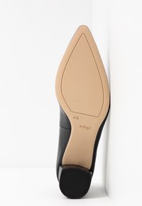 Semelle de chaussure noire en cuir à bout pointu tournée vers le haut, dessous beige avec la taille 4½ et le logo "högl", appuyée contre une surface blanche.