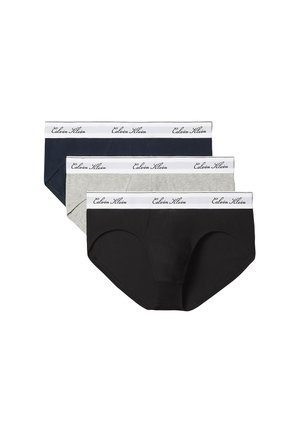 Muskuløs ung mand med krøllet hår står barfodet mod en ensfarvet baggrund iført beige Calvin Klein underbukser.