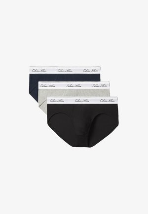 Muskelöser junger Mann mit lockigem Haar steht barfuß vor einem schlichten Hintergrund und trägt beige Calvin Klein Slips.
