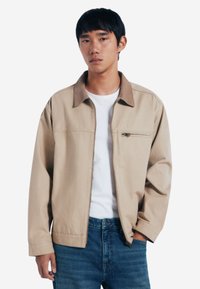 Giacca zip-up beige con colletto marrone a contrasto, dotata di tasca sul petto e vestibilità rilassata, abbinata a jeans di denim blu.