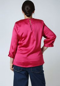 Woman with dark hair in a bun wearing a shiny fuchsia blouse with pearl buttons on the back and dark blue denim pants, standing with one hand on hip.