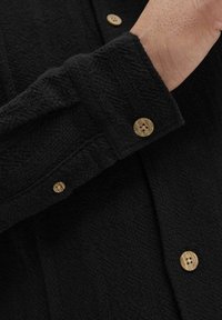 Manga de camisa negra texturada con botones de madera clara, que presenta un patrón sutil y un diseño de corte holgado. Vista cercana que muestra los detalles de la tela.