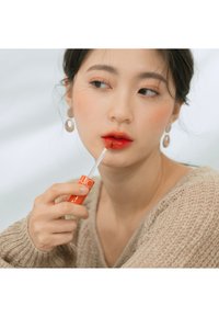 Brillo labial rojo en un tubo naranja, aplicado en los labios, sostenido por una persona que lleva un suéter de punto beige. Acabado preciso y brillante en los labios.