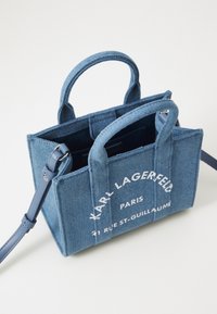 Sac en denim avec doubles poignées supérieures, bandoulière réglable et impression du logo blanc. Présente une forme structurée et une couleur indigo profonde.