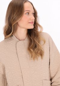 Beige steppelt kabát virágmintával, magas gallérral és oldalsó zsebekkel. Puha tapintású és laza fit, alkalmas hétköznapi viseletre.