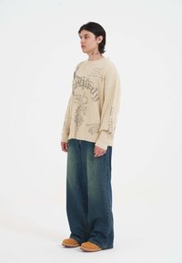 Beige gestreifter Pullover mit grafischen Details, kombiniert mit lässigen dunkelblauen Jeans und hellbraunen Slippern.