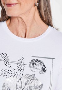 Weißes Baumwoll-T-Shirt mit Rundhalsausschnitt, versehen mit schwarz-silbernen botanischen Druckmustern und grafischen Akzenten. Sanfte Textur.