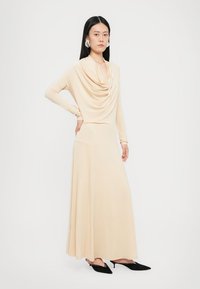 Beige pitkähihaineen mekko, jossa on laskostettu kaula-aukko ja virtaava helma. Valmistettu sileästä kankaasta, yhdistetty mustiin kärjet kärjellisiin korkokenkiin.