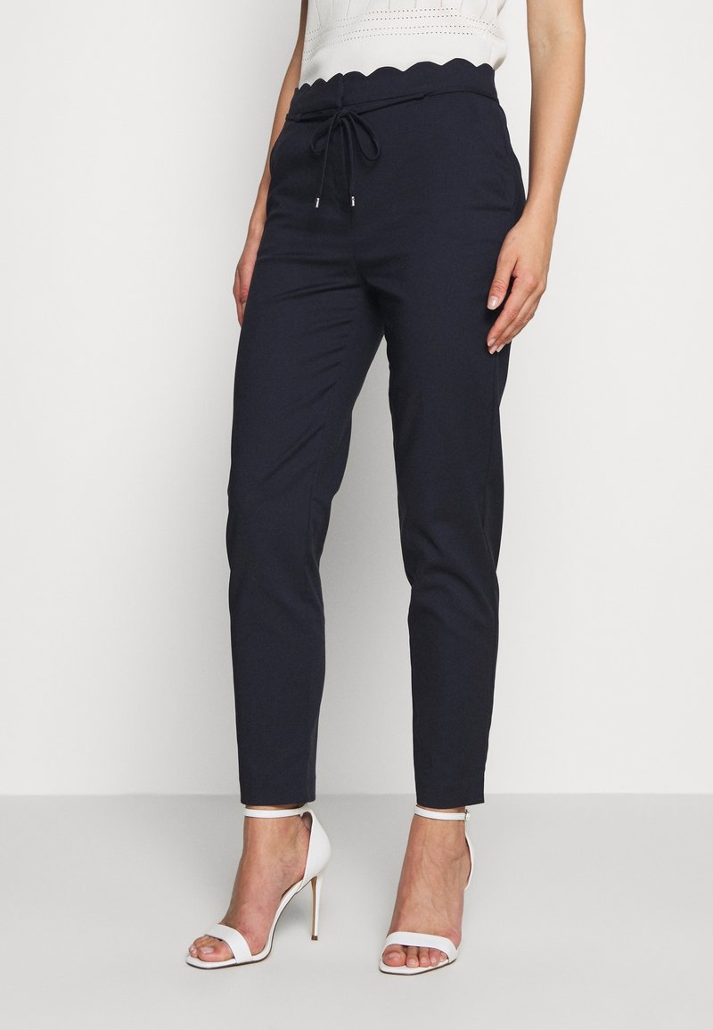 Femme portant un pantalon noir fuselé avec une taille festonnée et des sandales blanches à talons hauts, debout devant un fond blanc.