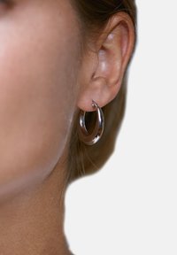 Hõbedane rõngaskõrvarõngas, sile ja läikiva viimistlusega, ümara kujuga, kinnitatud kõrvapadjale, minimaalne riistvara detail nähtav.