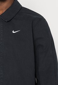 Černá bavlněná bunda s límečkem, na hrudi s bílým logem Nike a jemnou texturou viditelnou v látce.