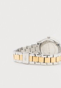 Montre-bracelet en acier inoxydable argenté et doré avec un bracelet à maillons. Elle présente un boîtier rond, un fermoir gravé du logo et un fond de boîtier transparent.