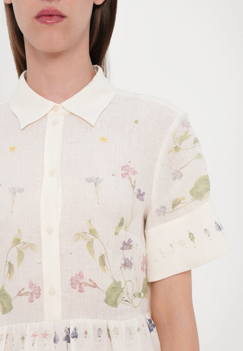 Robe en lin à manches courtes, de couleur crème, ornée d'imprimés floraux en violet, rose et vert, détaillée par de petites touches et une fermeture à boutons.