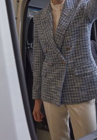 Modrý a bílý tvídový dvouřadý blejzr se zlatými knoflíky. Měkká textura, špičaté klopy, kombinovaný s krémovými kalhotami.