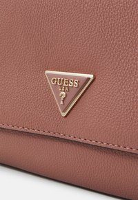 Rosa texturerad läderhandväska med en gyllene triangelformad Guess-logga och kontrasterande sömmar längs kanterna.