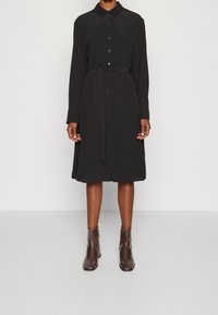 Abito camicia lungo nero a maniche lunghe con colletto, chiusura frontale con bottoni, cintura in vita e silhouette dritta, abbinato a stivaletti beige.