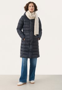 Navy pufferjas met verticale gestikte patronen, vo pockets en een capuchon. Gestyled met een crèmekleurige sjaal, gecombineerd met blauwe flared jeans en witte sneakers.