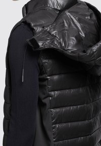 Czarna pikowana kamizelka z błyszczącym wykończeniem, z kapturem, bocznymi panelami z granatowej prążkowanej tkaniny oraz suwakiem z ozdobnym uchwytem.