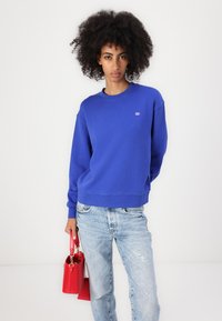 Sudadera de algodón azul con cuello redondo y puños de canalé, combinada con jeans de lavado claro y un bolso rojo. Detalle del logo simple visible.