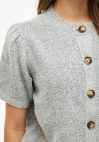 Gaiši pelēks īsroku džersija kardigans ar teksturētu apdari un pieciem brūniem pogām priekšpusē. Ir apaļš apkaklis un pūstie piedurknes.