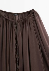 Blouse marron ample avec col élastiqué froncé et fermeture à nouer à l’avant, en tissu doux et léger.