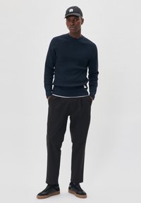 Navy ribbed sweater, black trousers, and black sneakers with gum soles. Black cap with a front patch. Casual, modern fit.