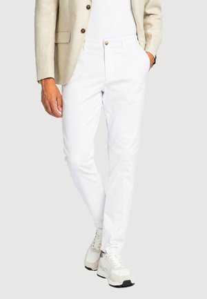 Uomo che indossa pantaloni bianchi, blazer beige, camicia bianca e scarpe da ginnastica bianche, in piedi con una mano in tasca e una mano rilassata lungo il fianco.