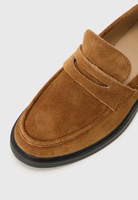 Mocassin en suédine brun avec un bout arrondi et un détail de sangle décorative, reposant sur un fond clair.