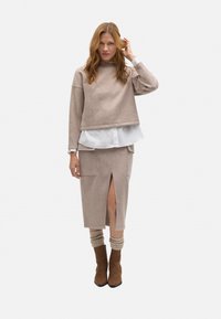 Sudadera recortada beige sobre una camiseta blanca, combinada con una falda midi beige con una abertura frontal y bolsillos laterales. Botas de ante marrón hasta el tobillo.
