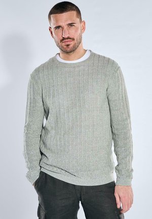 Mann mit kurzem Haar und Bart, der einen hellgrauen gerippten Pullover und dunkle Hosen trägt, steht vor einem einfarbigen hellen Hintergrund.