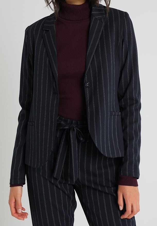 Femme portant un blazer bleu marine à fines rayures et un pantalon assorti avec une ceinture nouée, associé à un pull col roulé bordeaux.