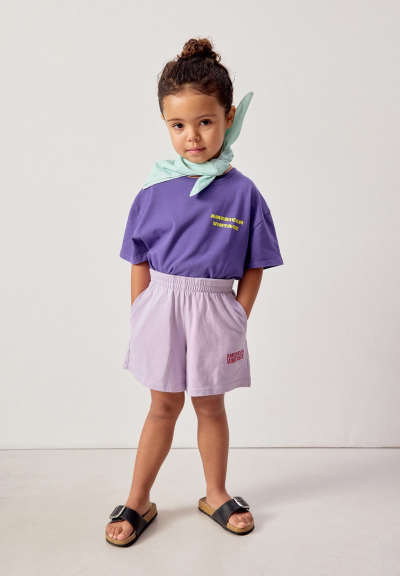 Jeune enfant aux cheveux bouclés attachés, portant un t-shirt violet "American Vintage", un short violet clair, des sandales noires et une écharpe vert menthe.