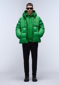Giacca a vento verde con cappuccio, tasche con zip e patch del logo, indossata sopra una felpa verde. Abbinata a pantaloni neri e stivali.