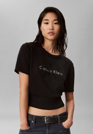 Młoda kobieta o mokrych, ciemnych włosach nosi czarny krótki sweter Calvin Klein z krótkim rękawem i ciemne dżinsy z czarnym paskiem. Ręce w kieszeniach.